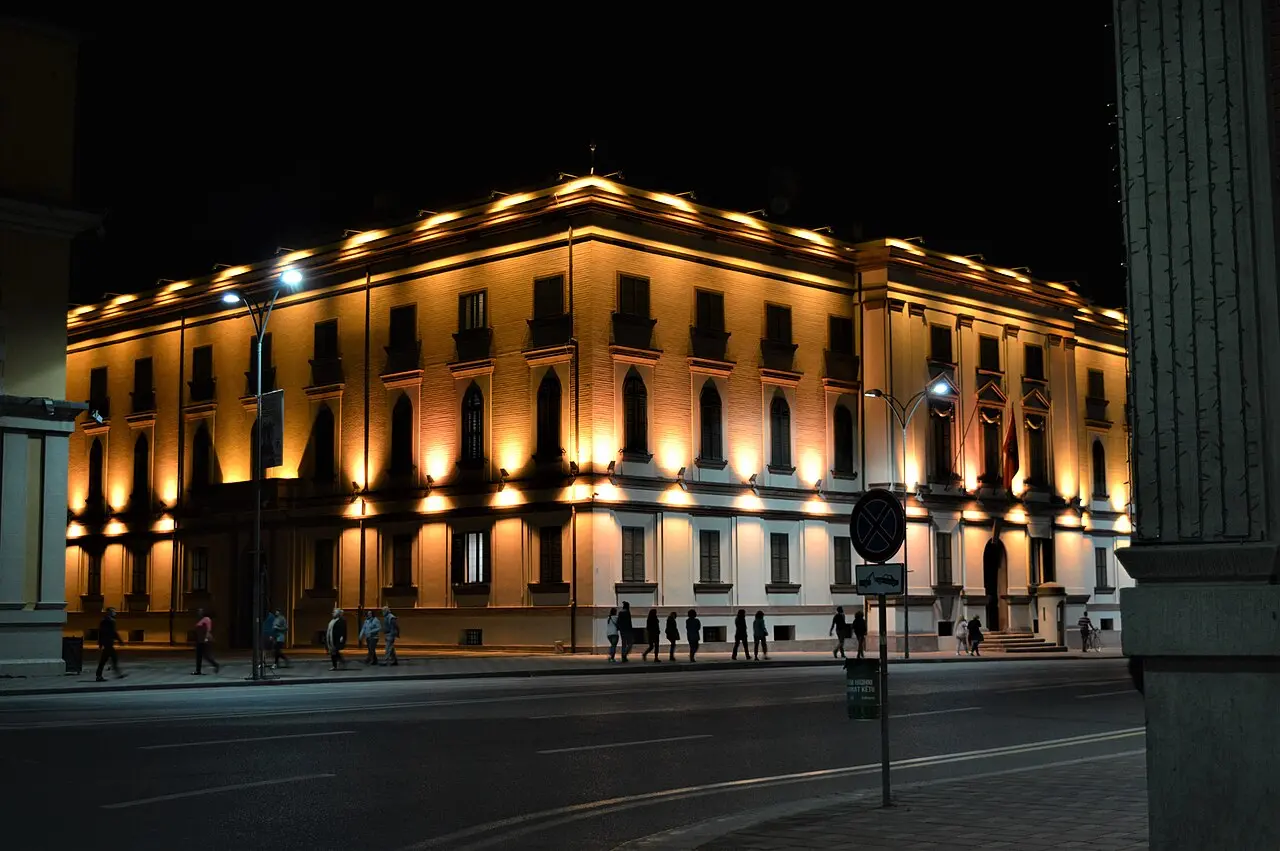 A grand illuminated building lit gold at night near the Tirana boulevard, with people walking past