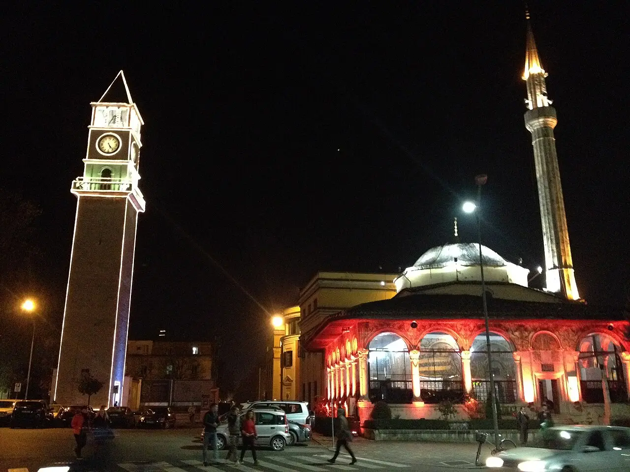 The lit Clock Tower and the Et'hem Bey Mosque at night in central Tirana, with people crossing the street