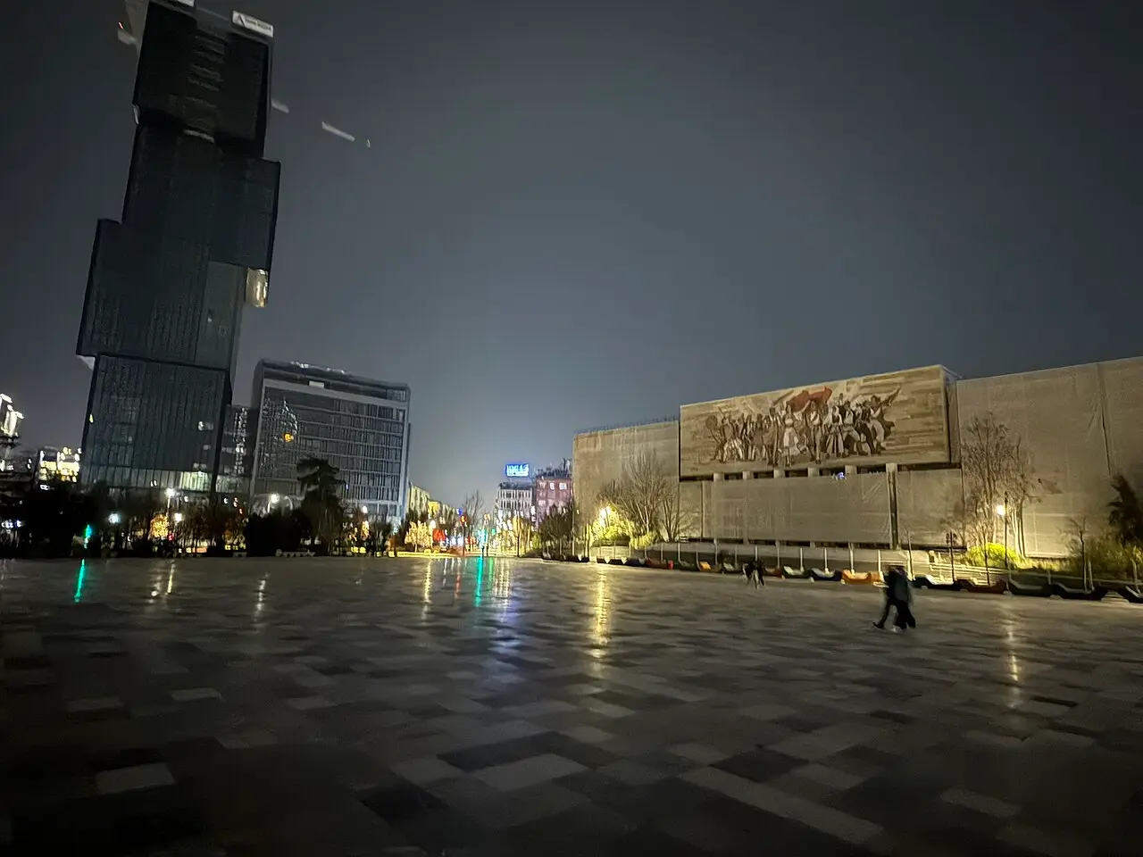 People crossing the wet, lamplit Skanderbeg Square in Tirana at night