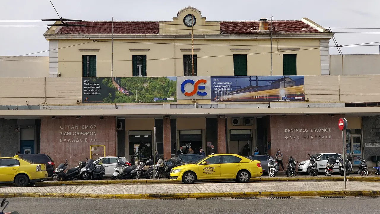 The cream facade of Athens Larissa railway station with a clock on the roof, an OSE sign and yellow taxis parked in front