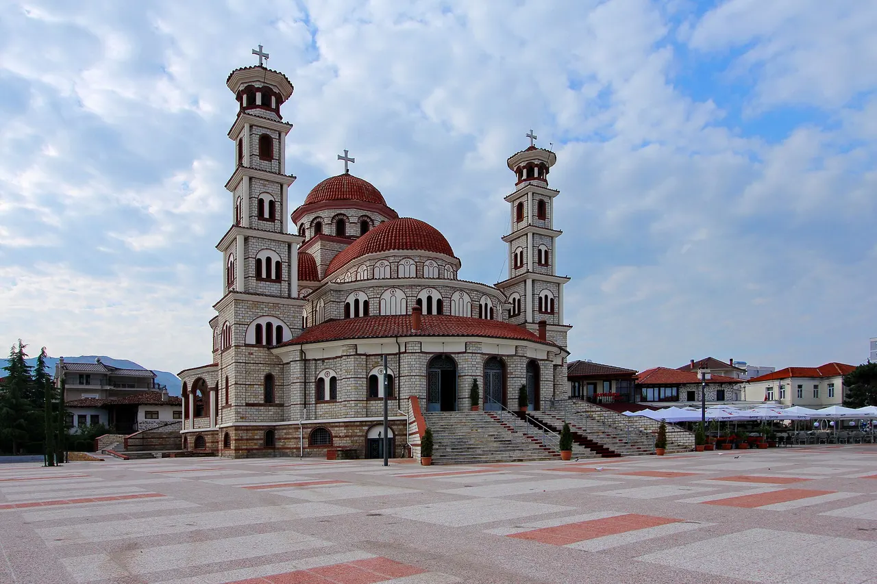 Korçë's Resurrection Cathedral with two bell towers and red domes on a wide paved square under a cloudy sky
