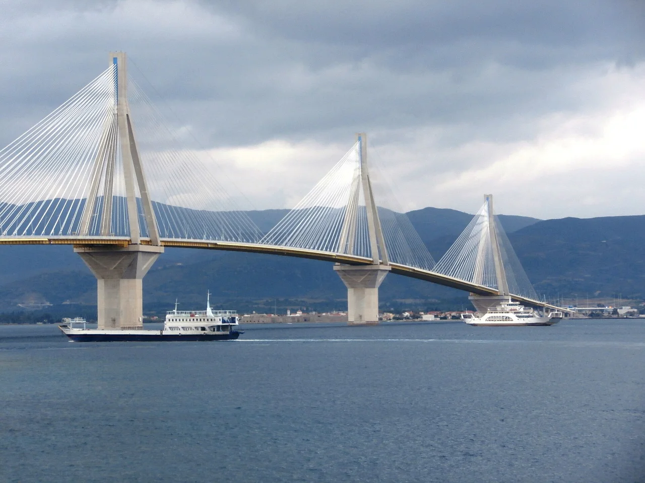 The cable-stayed Rio-Antirrio bridge over the Gulf of Corinth under grey cloud, two car ferries crossing beneath its pylons with mountains behind