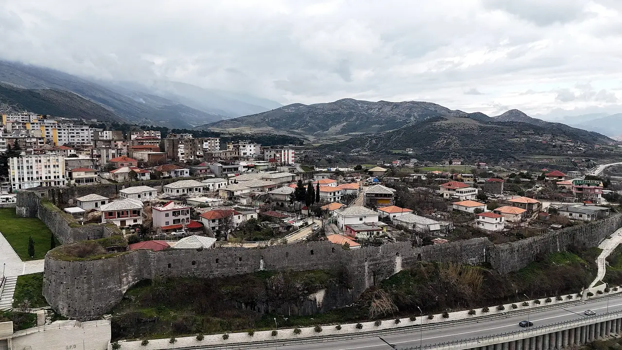 Aerial view of Tepelenë with its long grey fortress walls above the highway, houses inside the walls and green hills under low cloud behind
