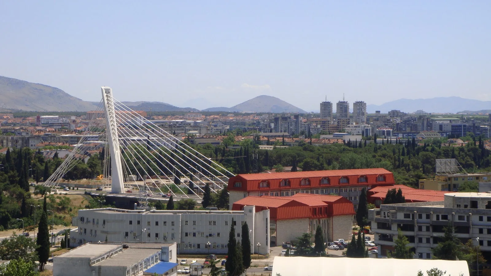 Podgorica seen from above, with the white cable-stayed Millennium Bridge, red-roofed buildings and mountains on the horizon