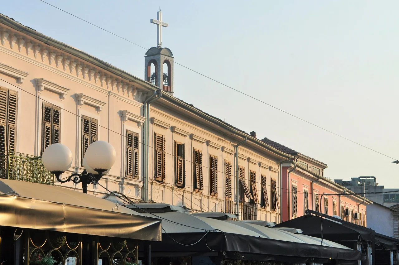 Pastel-coloured two-storey buildings with wooden shutters and café awnings along a pedestrian street in Shkodër