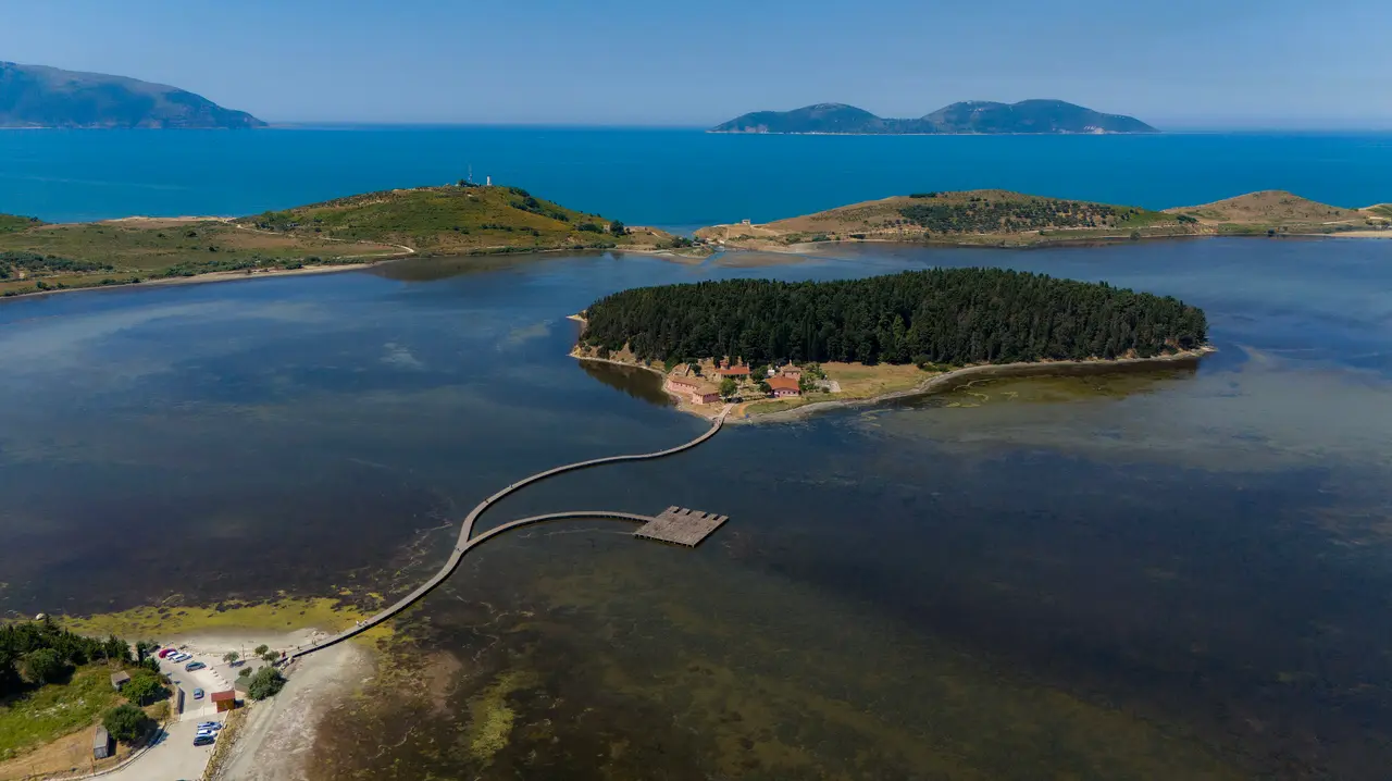 Aerial view of the wooden footbridge crossing the Narta lagoon to the pine-covered Zvernec island near Vlora, with the open sea and islands behind
