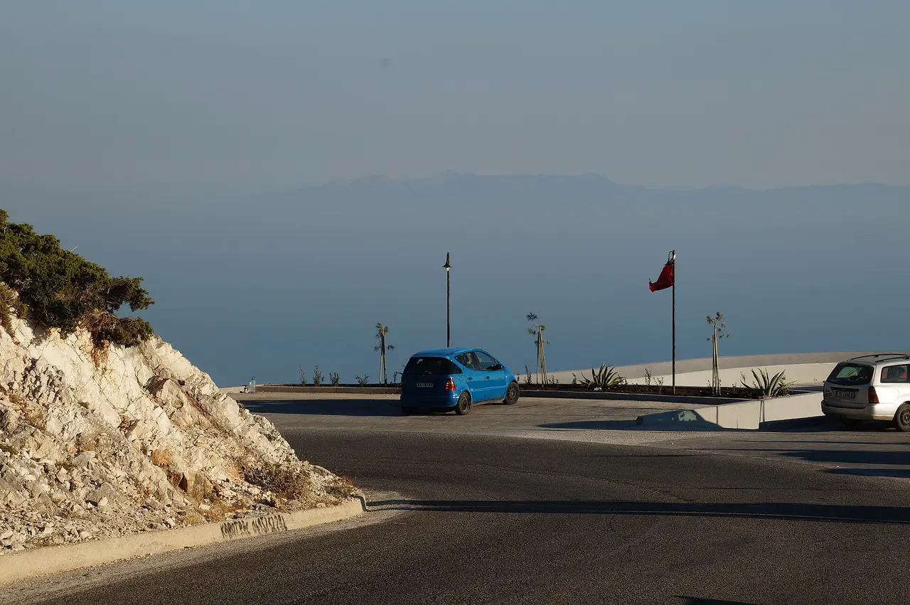 The car park at the Llogara viewpoint with an Albanian flag, parked cars and the hazy outline of Corfu across the water
