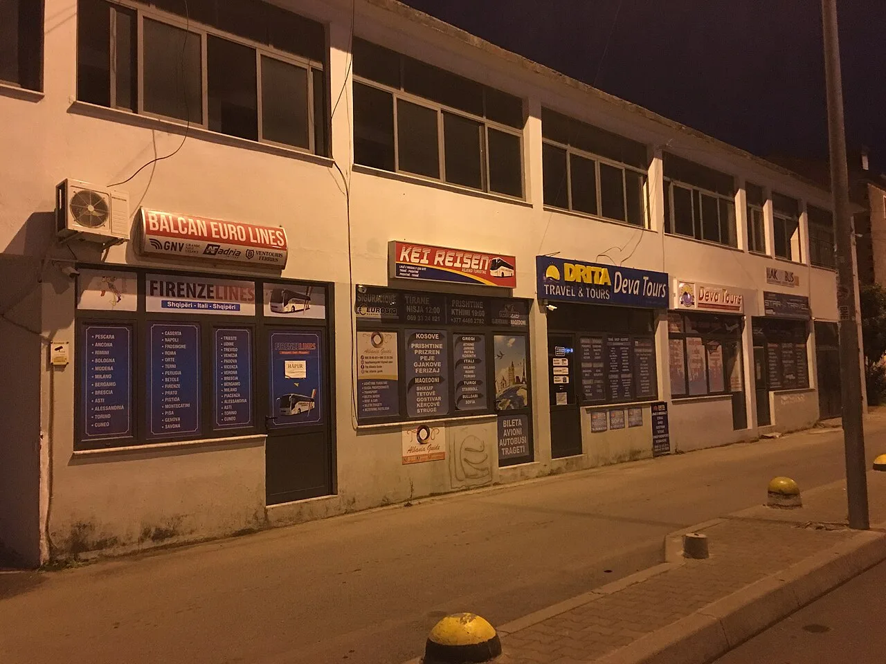A row of intercity bus and travel agency ticket offices in Tirana at dusk, advertising coach routes across Albania and abroad
