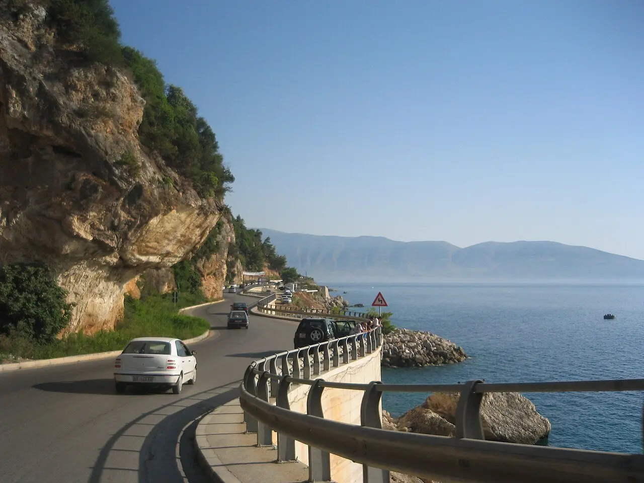 The coastal road cut into cliffs just south of Vlora, with cars winding along the rocky shoreline