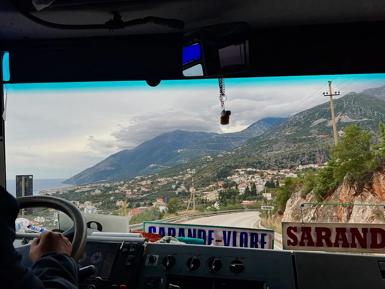 View through a bus windscreen descending toward a coastal town, with a hand-painted SARANDE-VLORE destination card taped to the dashboard