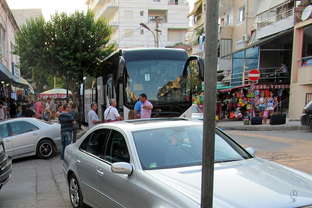 An intercity coach parked on a narrow street in Saranda with passengers and luggage on the pavement beside it