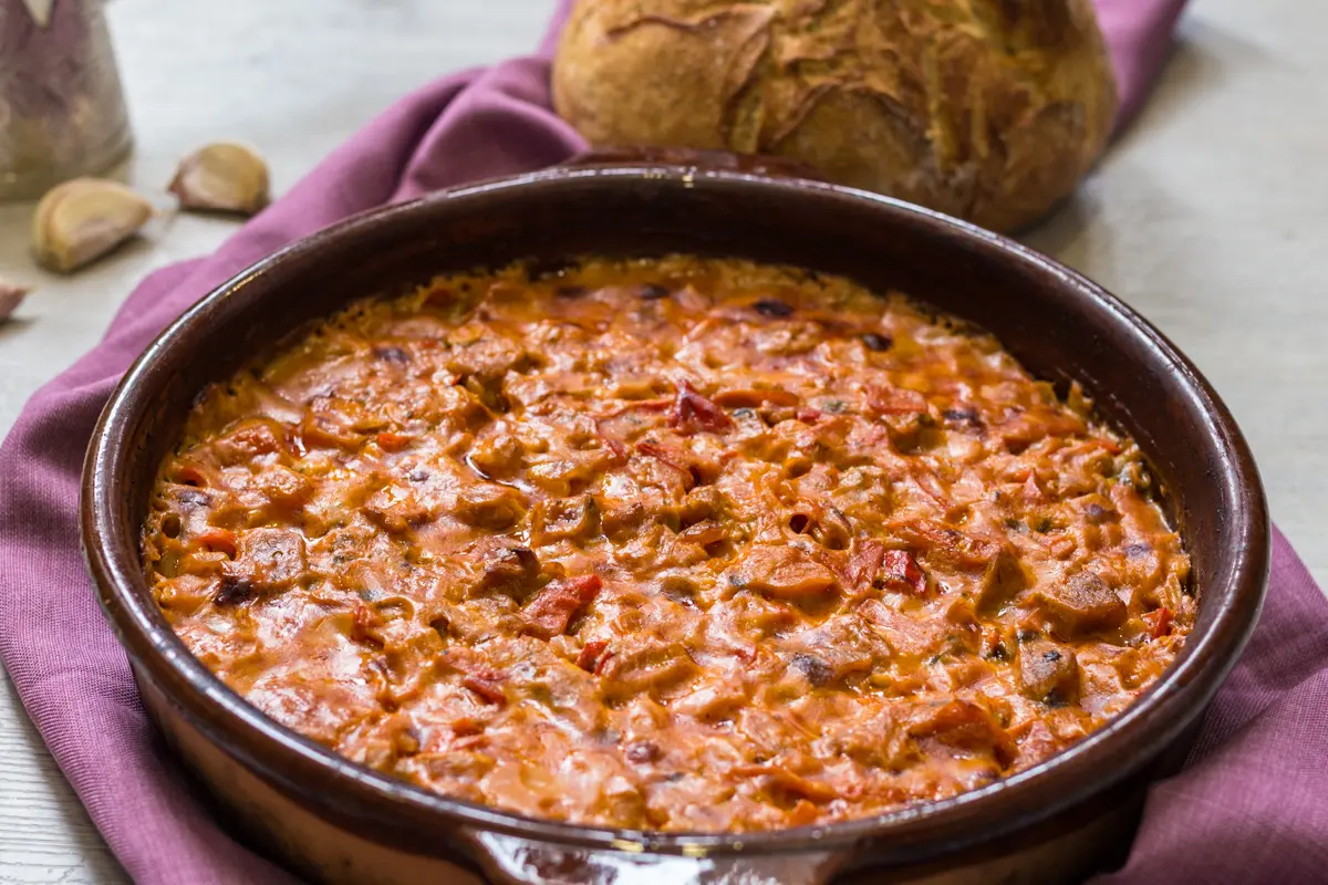 A clay dish of fërgesë, an Albanian bake of peppers, tomatoes and curd cheese, with a loaf of bread behind