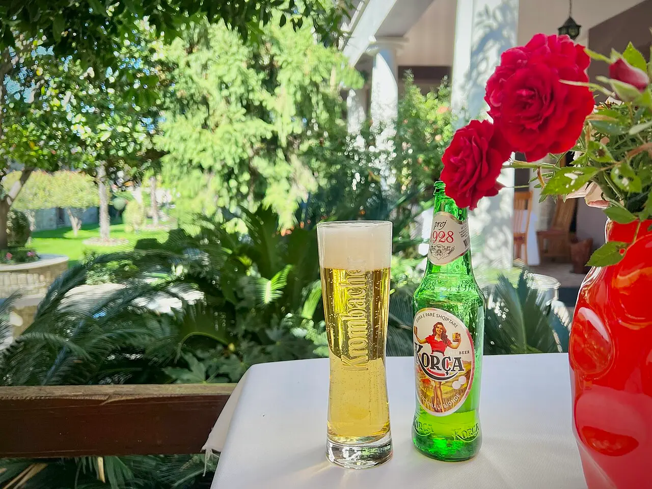 A green bottle of Korça beer marked 1928 and a poured glass on a sunny garden terrace
