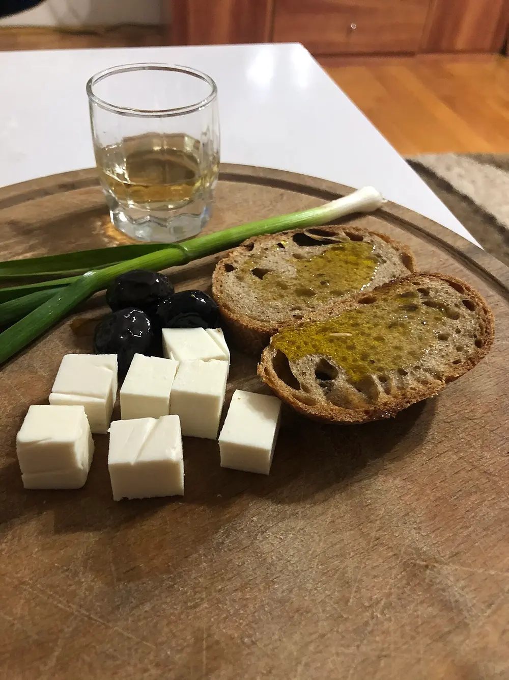 A wooden board with cubed white cheese, black olives, spring onions, bread with olive oil and a glass of raki