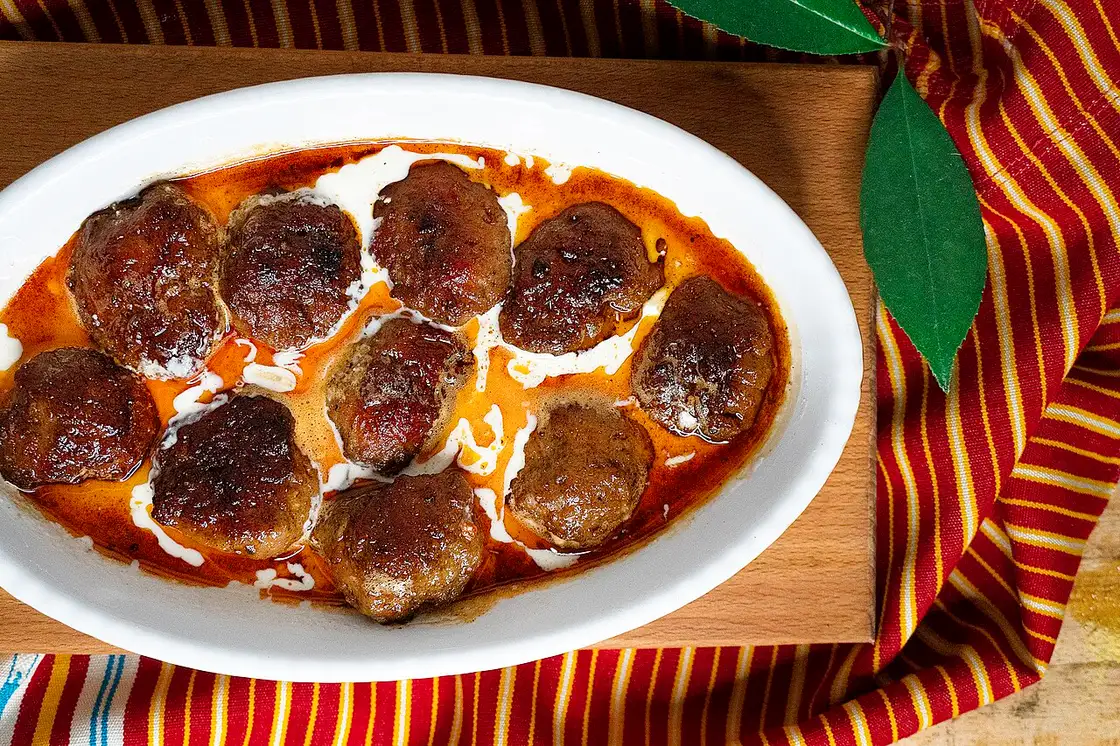 A white platter of Albanian qofte meatballs in a rich sauce on a striped cloth