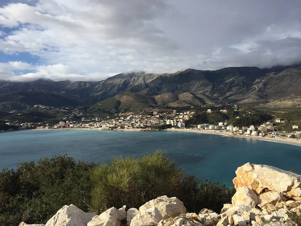Elevated view over Himara's bay and beach, with the town curving around the shore and the Ceraunian Mountains rising behind