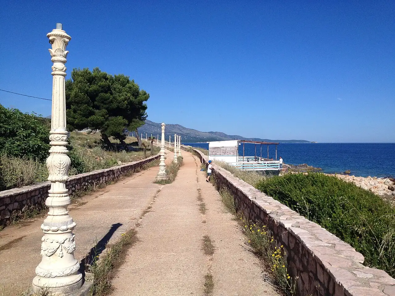 A seaside boardwalk in Ksamil leading towards the water, with a decorative lamppost, blue sky and the Ionian Sea