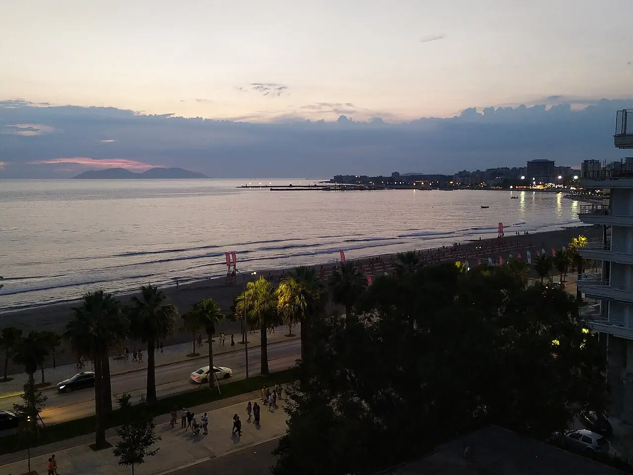 Sunset over Vlora bay with the lit seafront promenade, palm trees, beach and Sazan island on the horizon