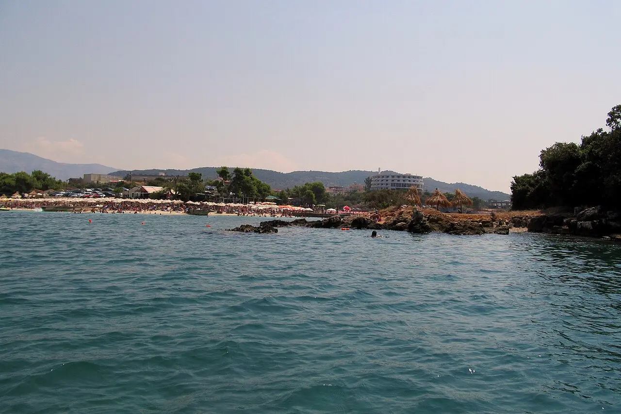 A wider view of a Ksamil bay with parasols along the shore and a large hotel on the wooded hillside behind