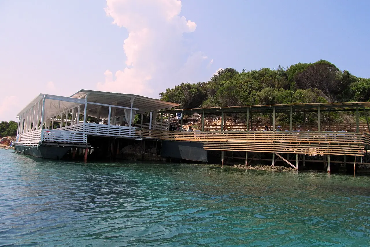 A wooden beach-bar platform built out over clear turquoise water beside a pine-covered islet at Ksamil