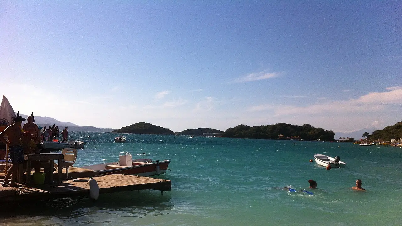 A turquoise bay at Ksamil with small offshore islets, a wooden jetty and swimmers in the shallow water