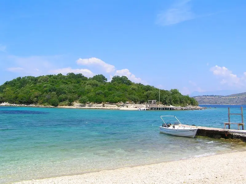 A small boat moored at a jetty in front of a wooded islet in the clear turquoise water off Ksamil