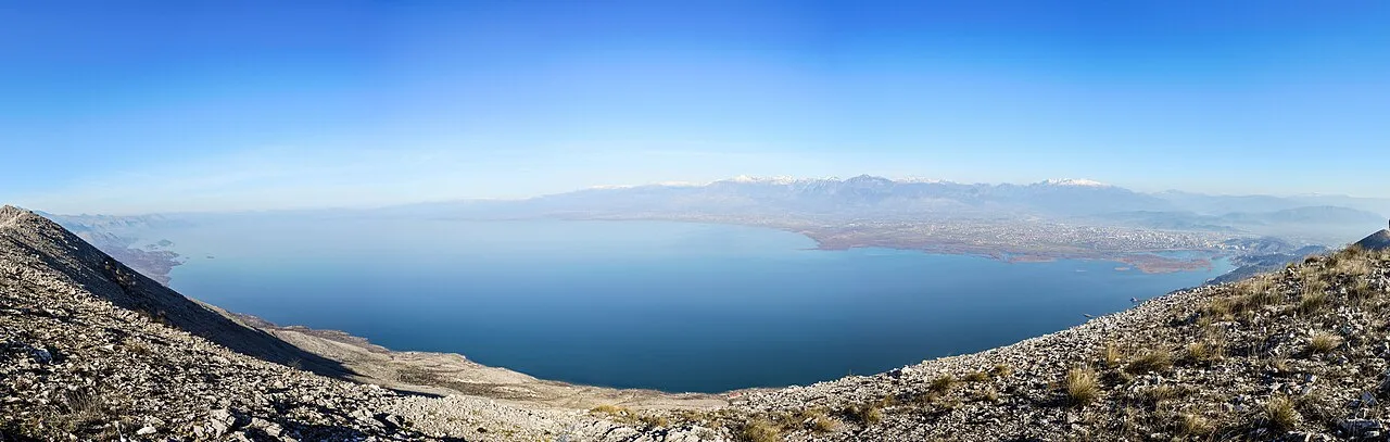 A wide panorama of Lake Shkodra with the city in the distance and the Albanian Alps rising behind it