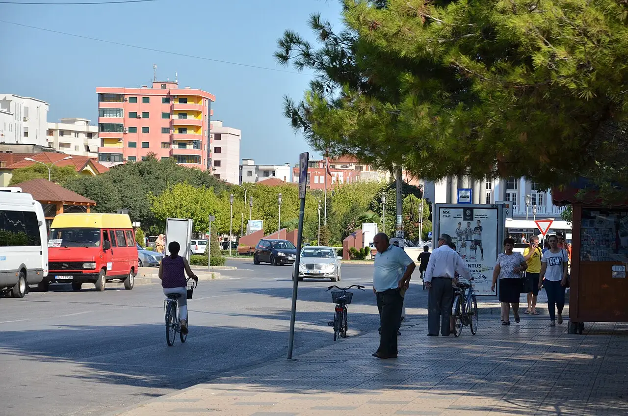 A tree-lined residential street on the northern edge of Shkodra with cyclists, minibuses and ordinary apartment blocks