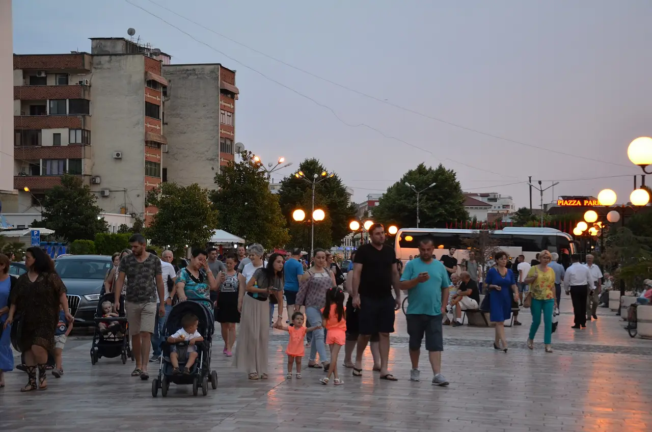 Families and groups walking the wide pedestrian promenade in Shkodra at dusk under street lamps